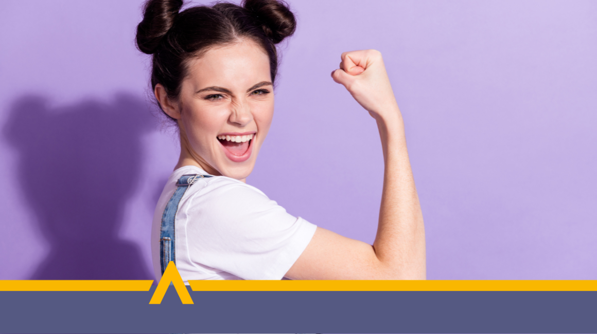 Kobieta w białej koszulce i ogrodniczkach uśmiecha się szeroko i pokazuje zaciśniętą pięść w geście zwycięstwa, na fioletowym tle.